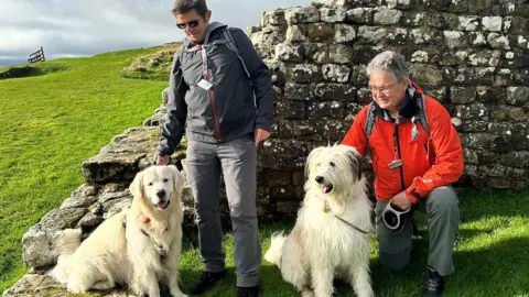 National Trust Tom Millmore (izquierda) está al lado del Sr. Darcy, un golden retriever. Junto a él, Charlie Wainwright está arrodillado al lado de Bear, un perro pastorgherd Myoritic. Los restos de la casa se pueden ver detrás con un área cubierta de hierba.