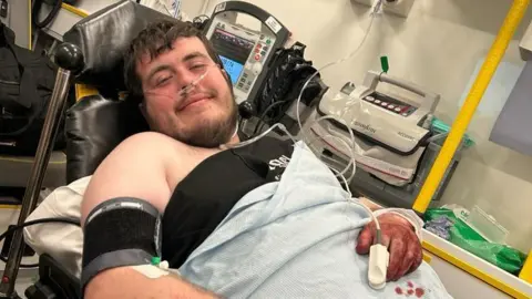 La fotografía de William Newbury de Newbury de William Newbury se dirigió a una cama de ambulancia. Lleva un tubo de oxígeno y tiene una lámina azul que cubre el cuerpo al cofre. Tiene cabello corto marrón y una barba marrón. Parece aturdido en la cámara con una sonrisa. Su mano derecha se encuentra en el estómago y está cubierta de sangre. 